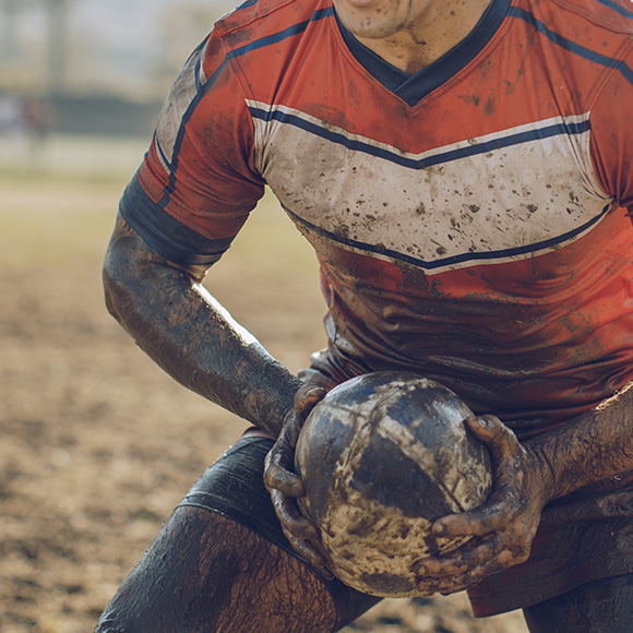 Muddy rugby player crouched with ball to promote leading rugby insurance broker Full-time Cover